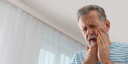 Älterer Mann mit Bart hält sich schmerzverzerrt die Wange, möglicherweise wegen Zahnschmerzen.