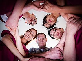 Fünf medizinische Fachkräfte bilden einen Kreis und schauen lächelnd nach unten in die Kamera, während sie sich gegenseitig an den Händen halten.