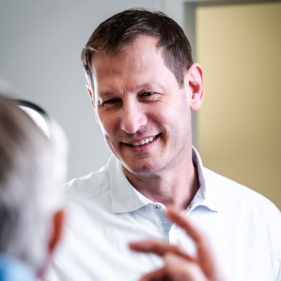 Zahnarzt im Gespräch mit einem älteren Patienten, lächelnd in freundlicher Atmosphäre.