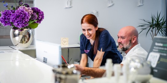 Mitarbeiterin in medizinischer Kleidung und ein Patient schauen gemeinsam auf einen Bildschirm an der Rezeption einer modernen Zahnarztpraxis.