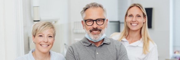 Drei freundlich lächelnde Personen, vermutlich ein Zahnarzt und zwei Praxismitarbeiterinnen, stehen in einer hellen, modernen Zahnarztpraxis.